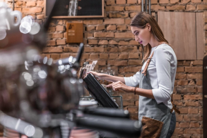 Jeune femme en tablier utilisant un écran de caisse tactile dans un café ou restaurant à l’ambiance chaleureuse avec mur de briques apparentes.